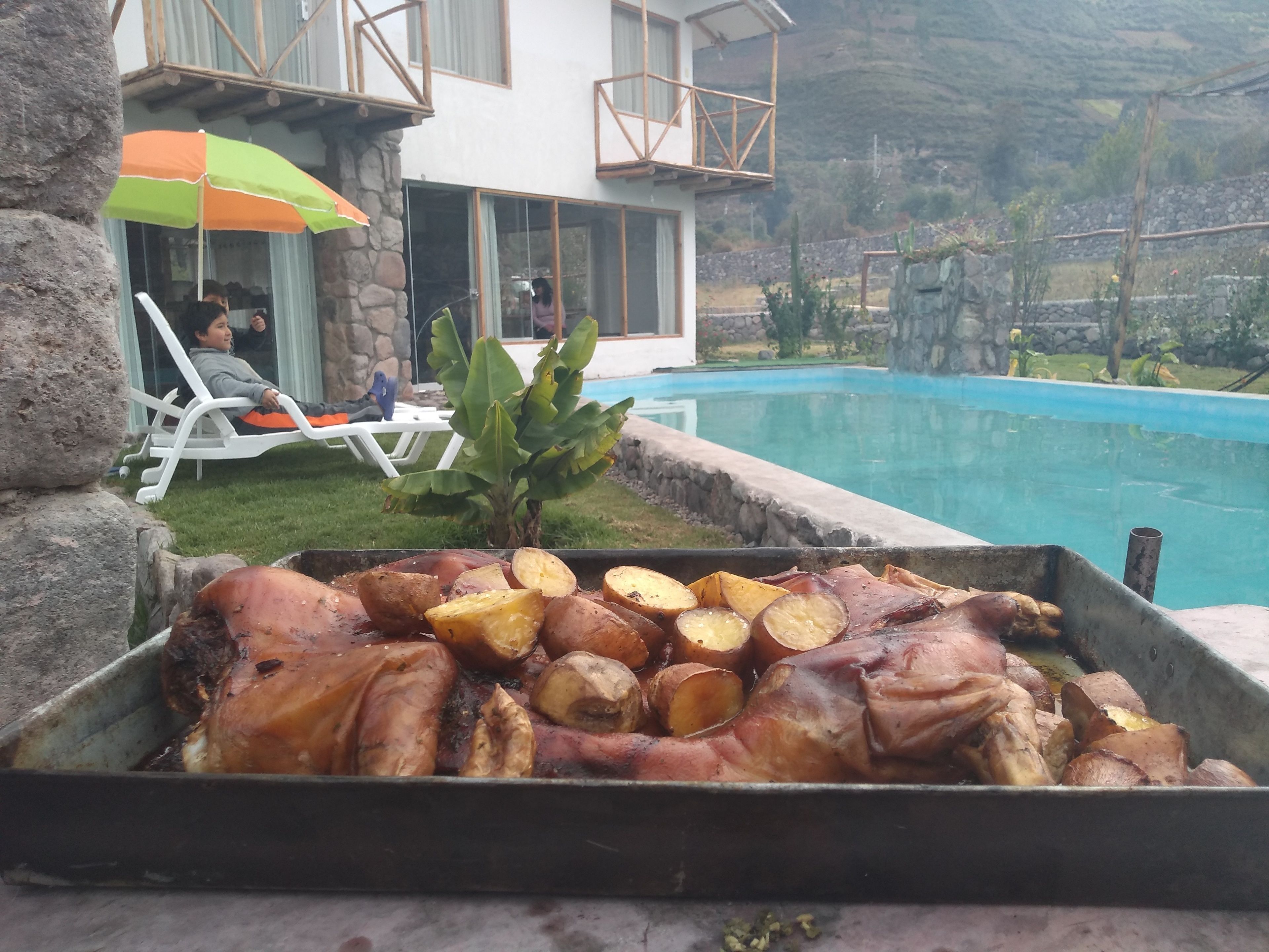 A poolside area with a barbecue grill containing roasted meat and potatoes, a stone wall, and a house with a balcony in the background.