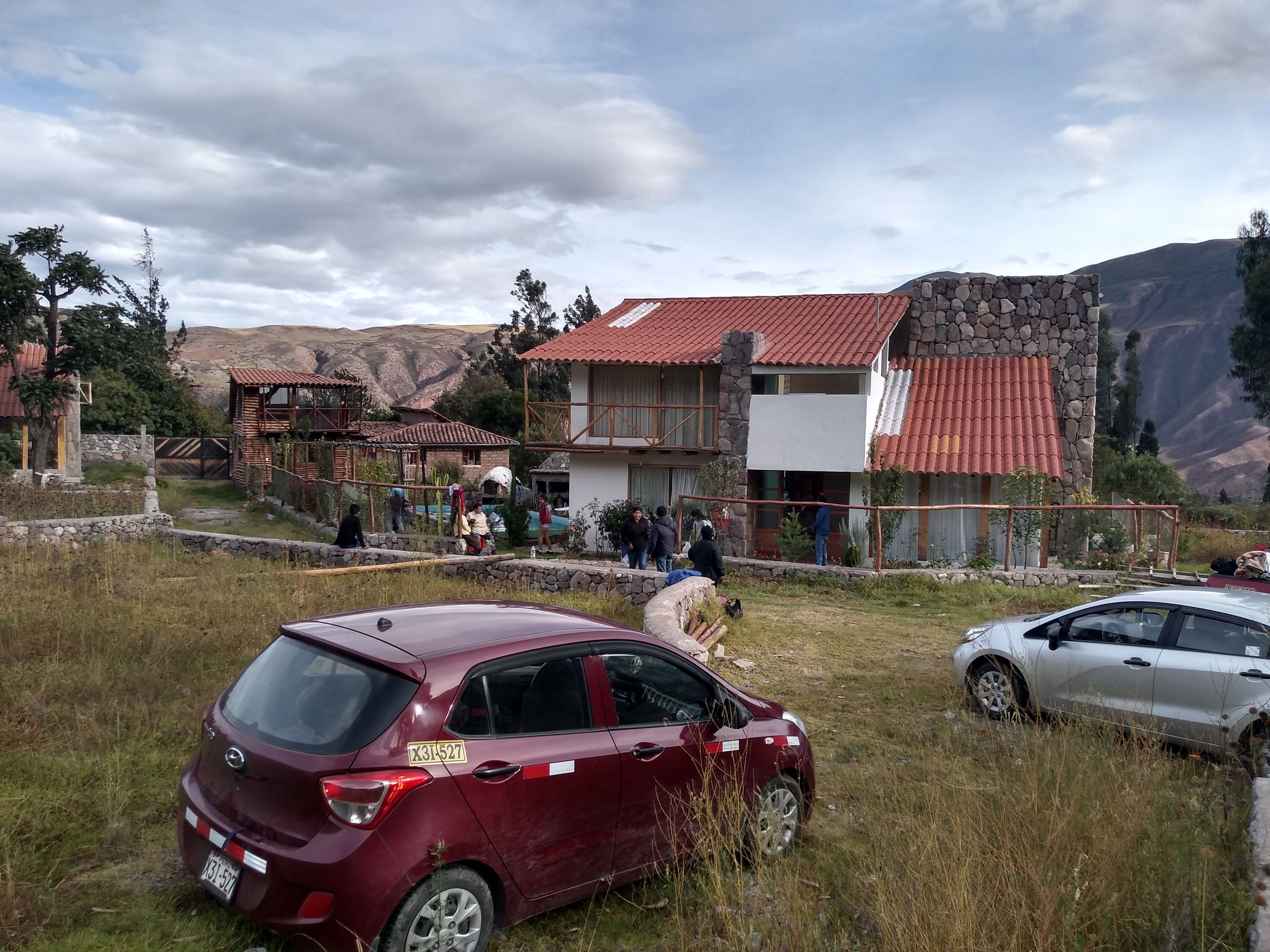 A rustic building with a stone facade and red-tiled roof, surrounded by a grassy area with parked cars.
