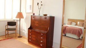 Une chambre avec une commode en bois, une chaise, une table de chevet et un lit recouvert d’une couverture rose.