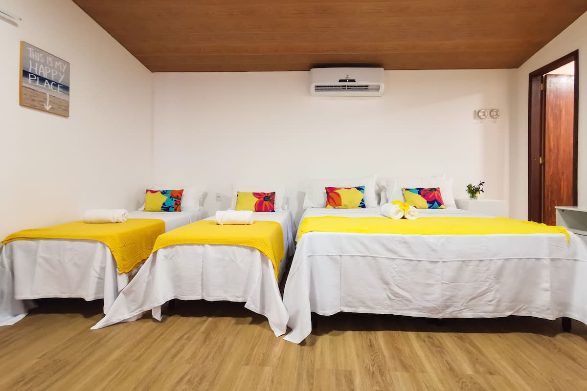 A hotel room with two beds, each with a yellow blanket and colorful pillows. There is a ceiling fan, an air conditioning unit, and a wooden floor.