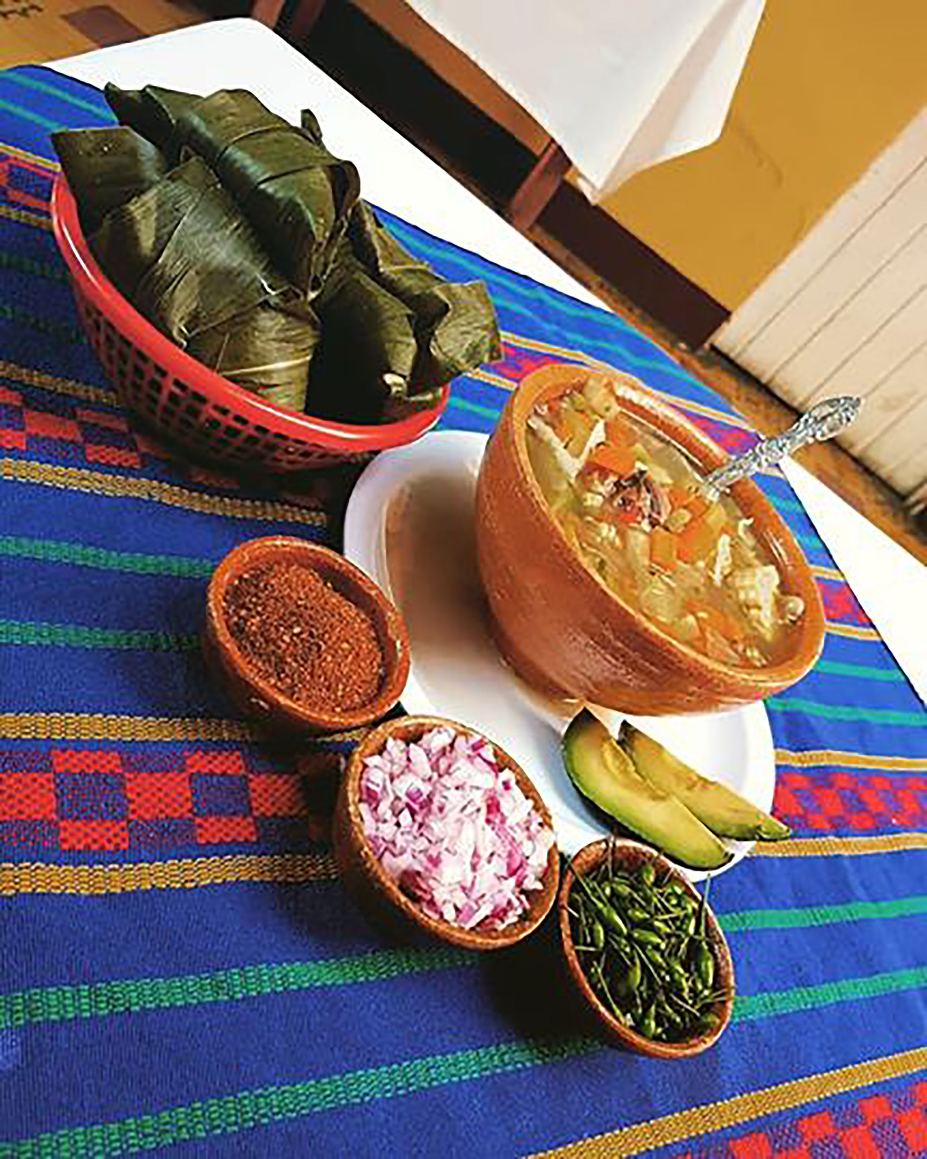 A table with a colorful woven tablecloth, a bowl of soup, a basket of tamales, a bowl of red onions, a bowl of green chilies, and an avocado.