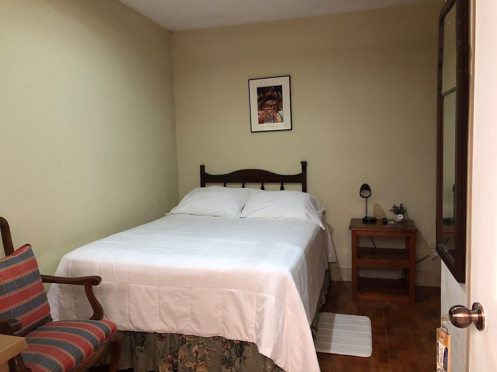 A bedroom with a bed, a chair, a small table with a lamp, and a picture on the wall.