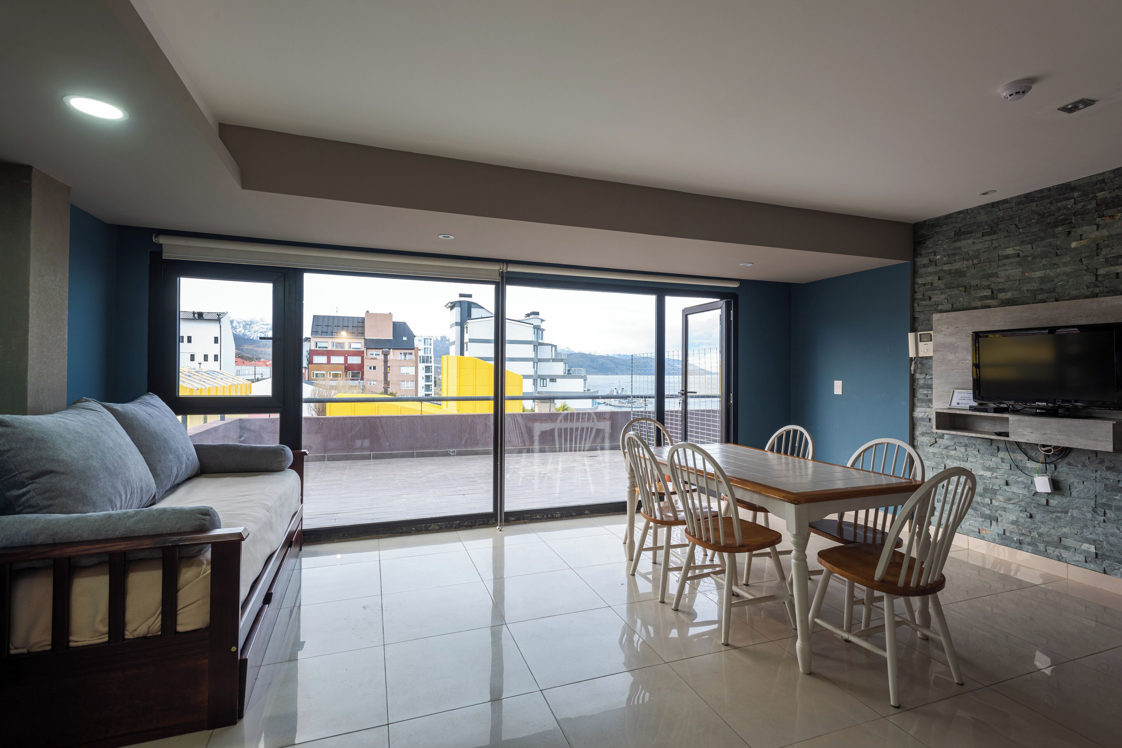 Ein modernes Wohnzimmer mit einer Couch, einem Esstisch, Stühlen, einem an einer Steinwand befestigten Fernseher und einem großen Fenster mit Blick auf die Stadt.