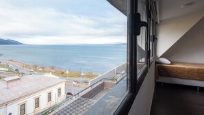 A room with a bed, a window with a view of a body of water and a distant shoreline, and a staircase leading to an outdoor area. - Gente del Sur - Rivadavia (Ushuaia)
