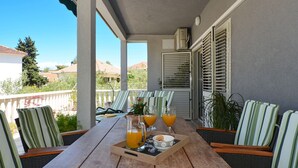 Een houten tafel met gestreepte stoelen, een dienblad met jus d'orange, en een schaal op een balkon met uitzicht op huizen en bomen.