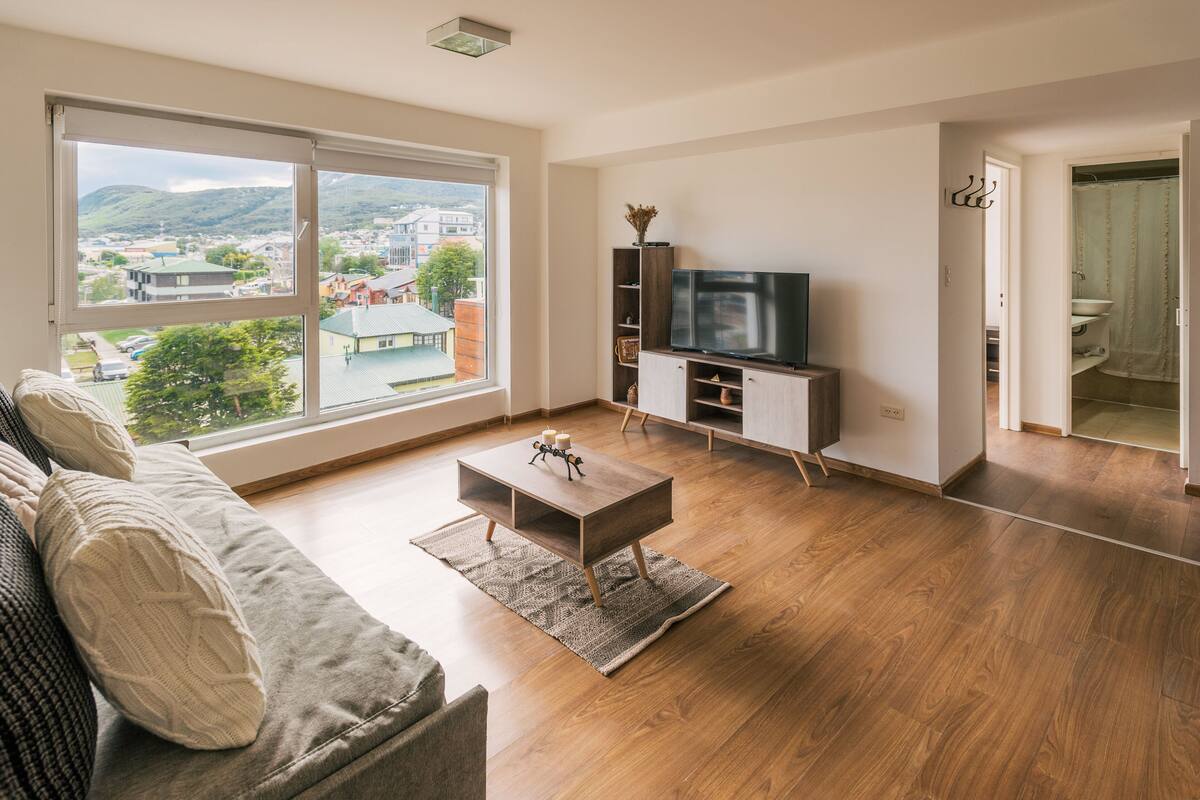 Ein modernes Wohnzimmer mit großem Fenster, Flachbildfernseher, Couchtisch aus Holz und einer Couch.