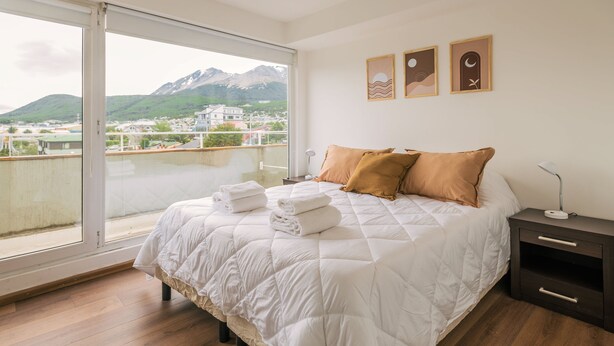 Un dormitorio con una cama grande, una mesita de noche, una lámpara y vistas a montañas a través de un ventanal amplio.