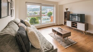 Una sala de estar moderna con un sofá, una mesa de centro, un televisor sobre un mueble bajo y un amplio ventanal con vistas a la ciudad.
