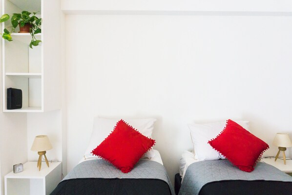 Habitación con dos camas, cada una con un almohadón rojo y uno blanco. Se ve un pequeño buró con una lámpara y un reloj a la izquierda. Una planta se distingue en la pared izquierda.