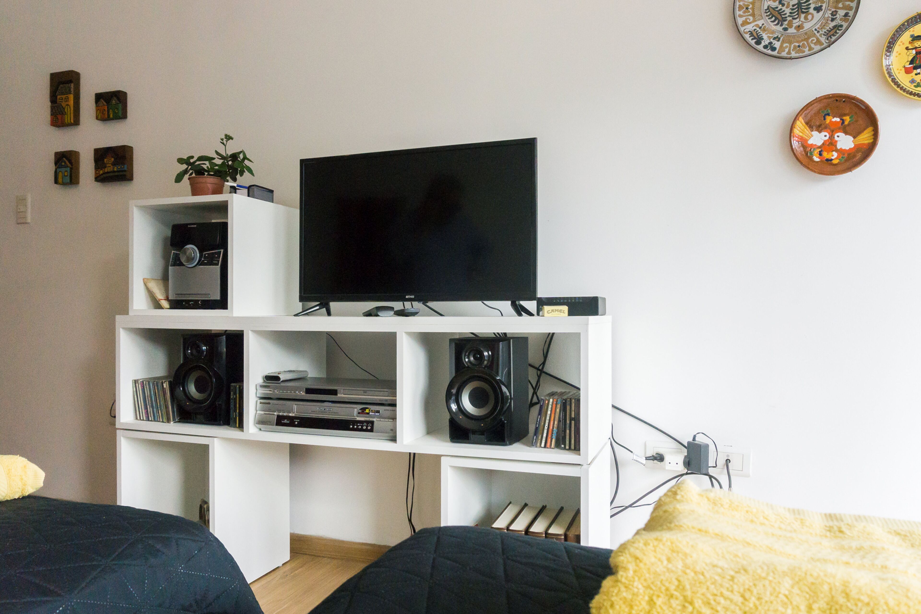 A living room with a TV on a white entertainment unit, surrounded by shelves with various electronic devices and books.