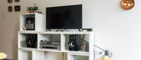 A living room with a TV on a white entertainment unit, surrounded by shelves with various electronic devices and books.