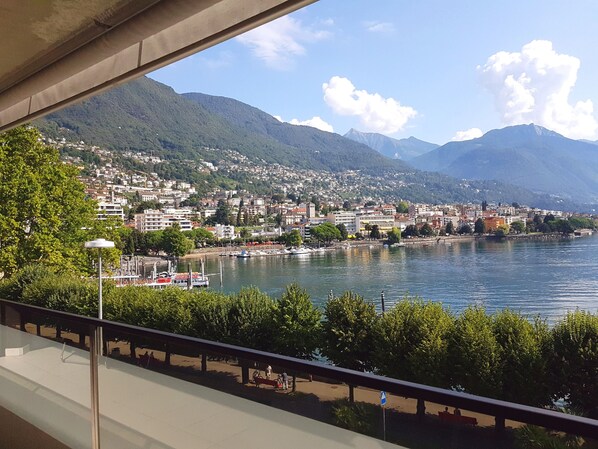 A panoramic view of a lakeside city with mountains in the background, a marina with boats, and a pedestrian walkway along the water.