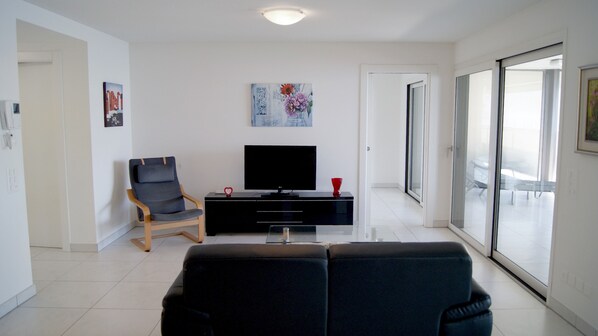 Una sala de estar moderna con un sofá negro, una silla de madera, un televisor sobre un mueble negro y un ventanal que da a un balcón.