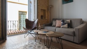 A living room with a grey sofa, a brown chair, a small round table, and a patterned rug. There is a balcony with a metal railing and a view of buildings. - Hommyhome Casa Assle 101 (Sevilla)
