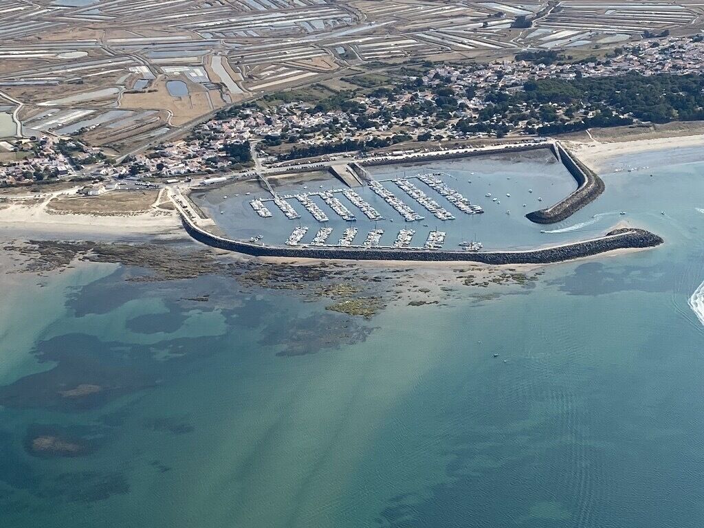 Vue aérienne d’un port de plaisance situé sur la côte, avec de nombreux quais et des bateaux, entouré de zones résidentielles et d’une plage.