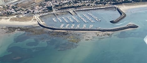 Vue aérienne d’un port de plaisance situé sur la côte, avec de nombreux quais et des bateaux, entouré de zones résidentielles et d’une plage.