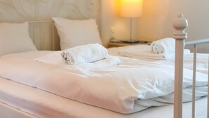 Een keurig opgemaakt bed met witte lakens en een opgerolde handdoek, een nachtkastje met een lamp en een hoofdbord met decoratief design.