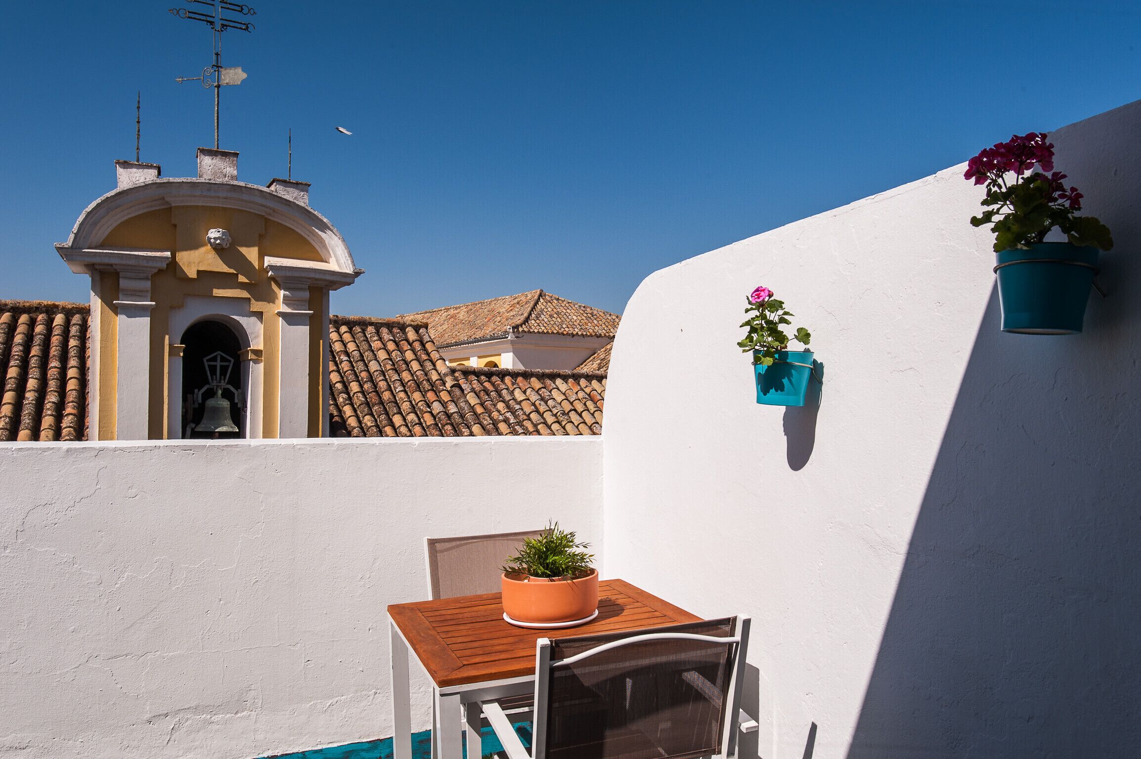 En lille terrasse med et træbord og stole, en potteplante og en hvid væg med en blå potte.