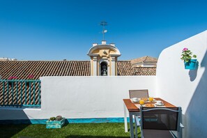 A terrace with a table set for breakfast, a white wall, and a building with a tiled roof in the background.