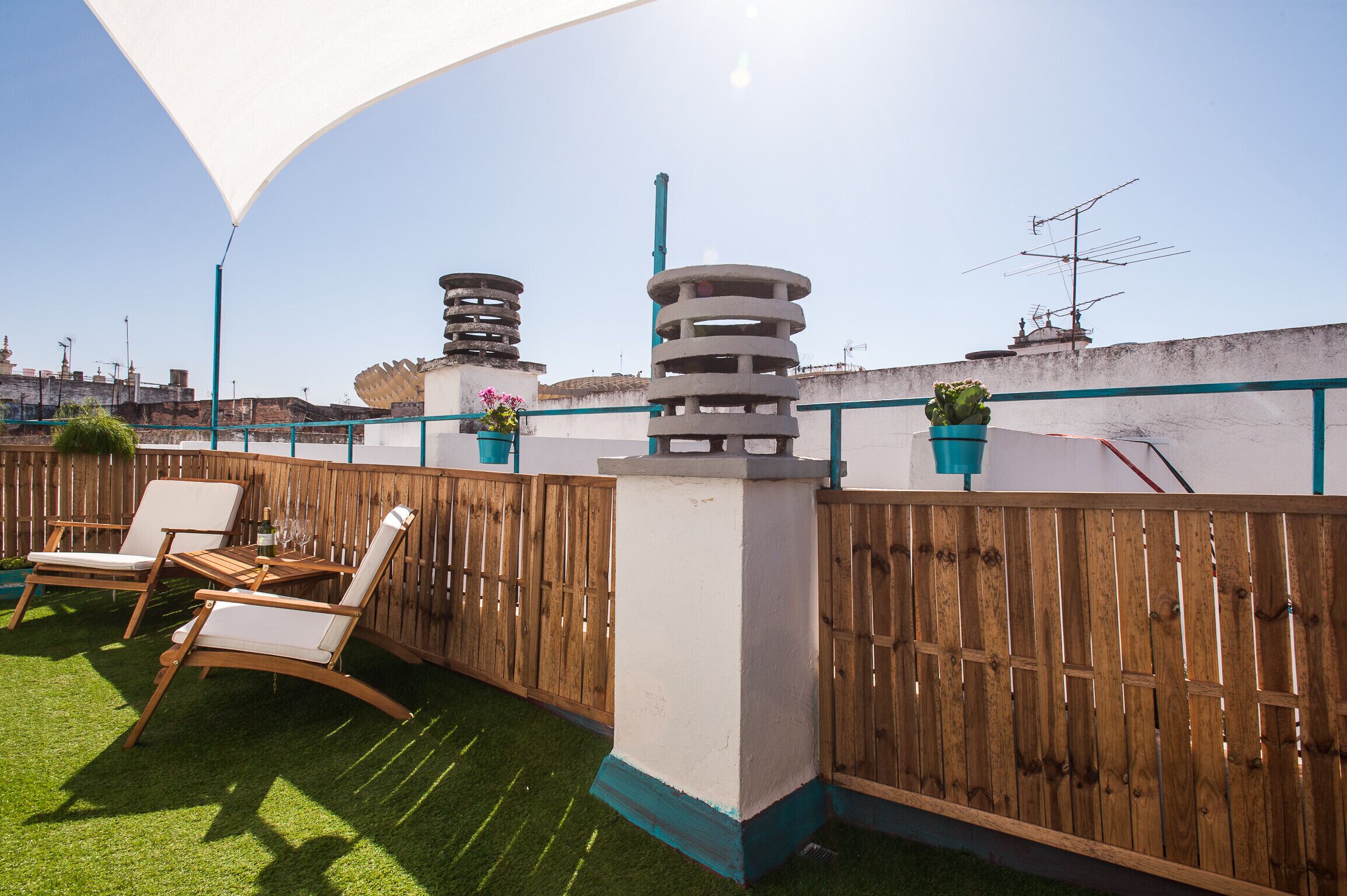 Eine Dachterrasse mit Holzlattenzaun, zwei Liegestühlen, einem kleinen Tisch und einem Blumentopf.