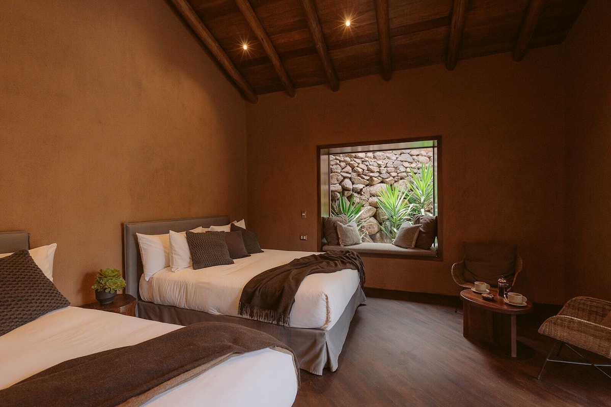 A bedroom with two beds, a window with a view of rocks and plants, a small table with cups and a teapot, and a chair.