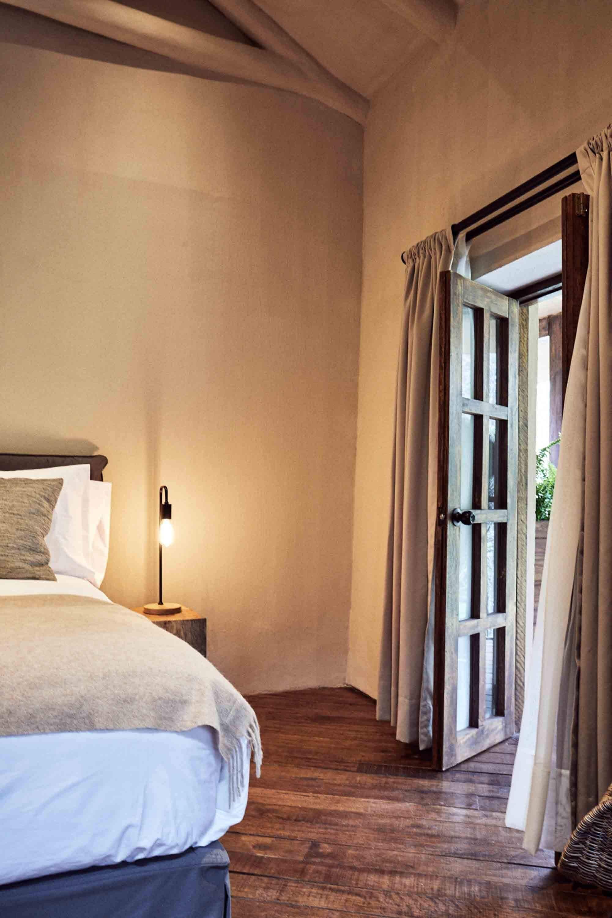 A bedroom with a wooden floor, a bed with white linens, a bedside lamp, and a door leading to a balcony with curtains.