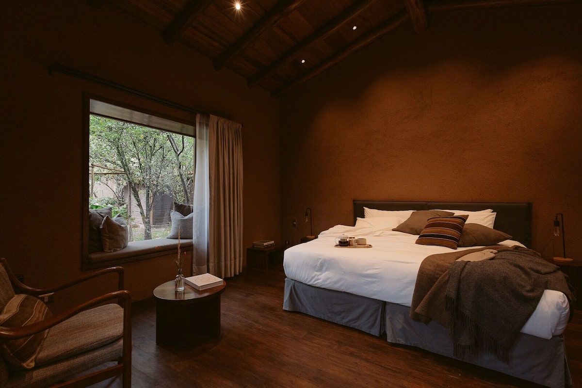 A bedroom with a large window, a bed with pillows, a bedside table, and a chair.