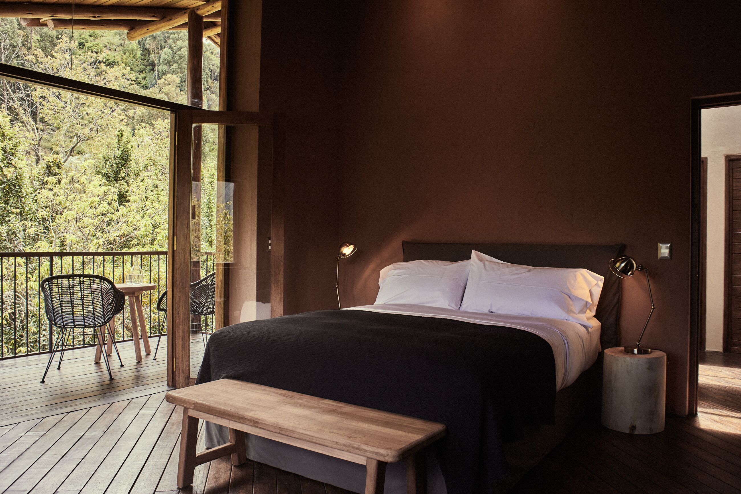 A bedroom with a large bed, a wooden bench, a small table, and a balcony with chairs.