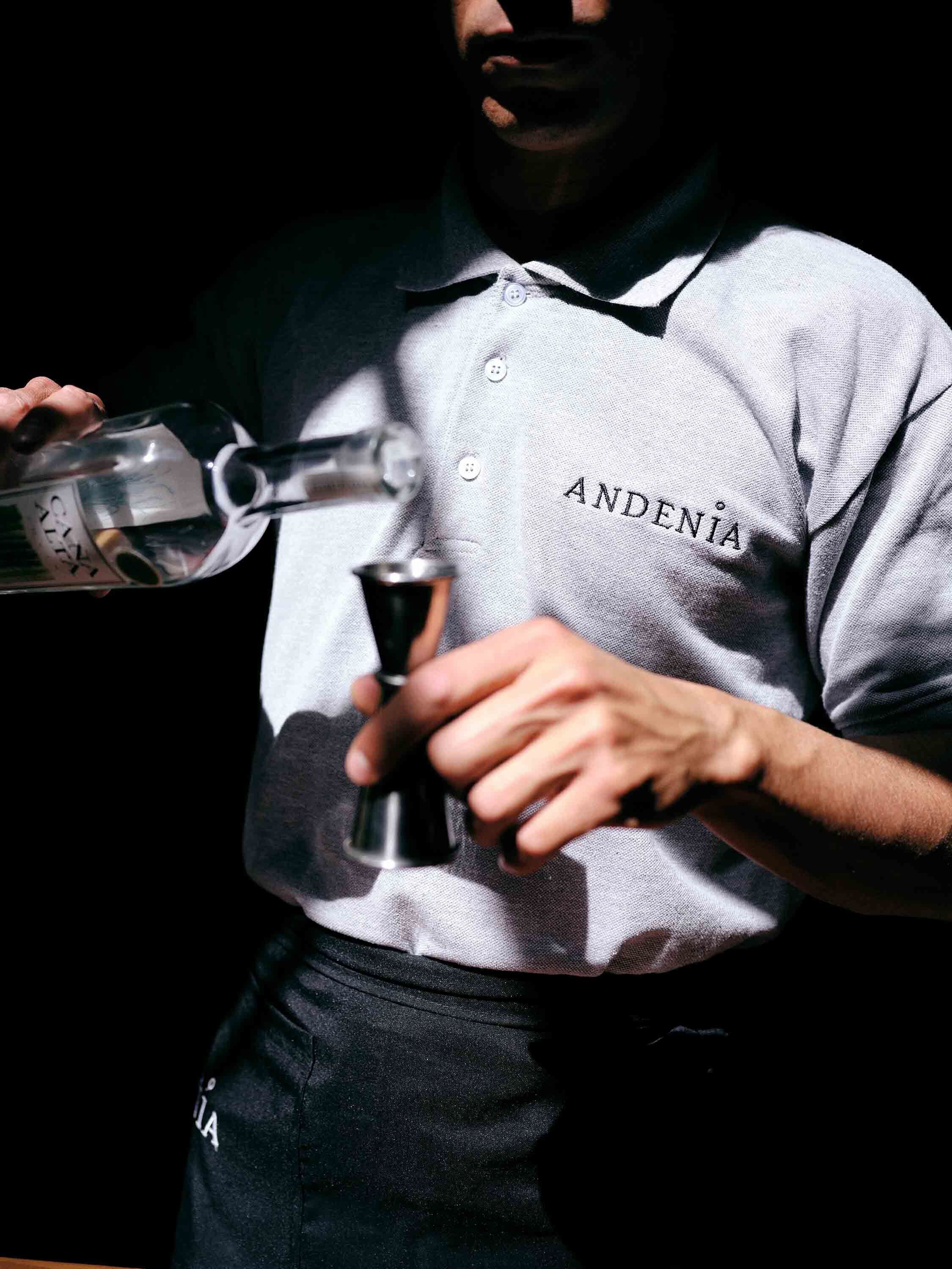 A person wearing a shirt with the word "ANDENIA" on it, holding a bottle and a glass.