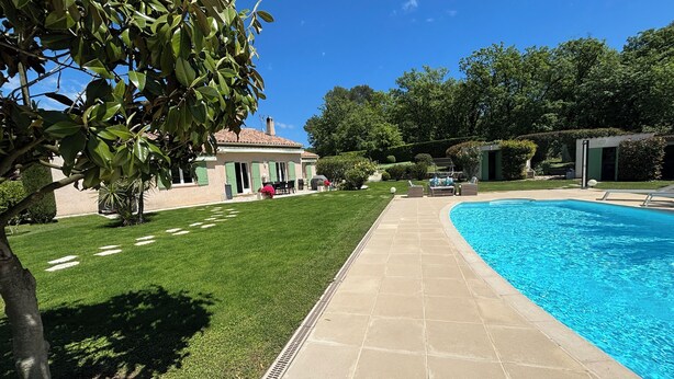 Une piscine avec une terrasse carrelée, une pelouse et un bâtiment doté d’une porte et de volets verts.