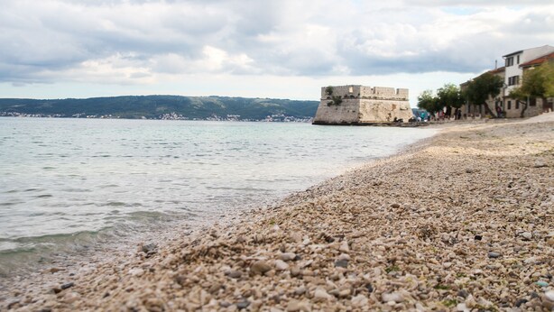 Ein Kiesstrand mit einer historischen Festung und Gebäuden entlang der Küste.