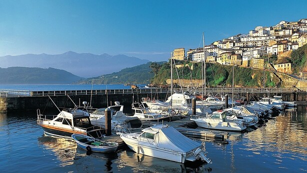 Un port de plaisance avec des bateaux amarrés, un pont et une ville perchée sur une colline en arrière-plan.