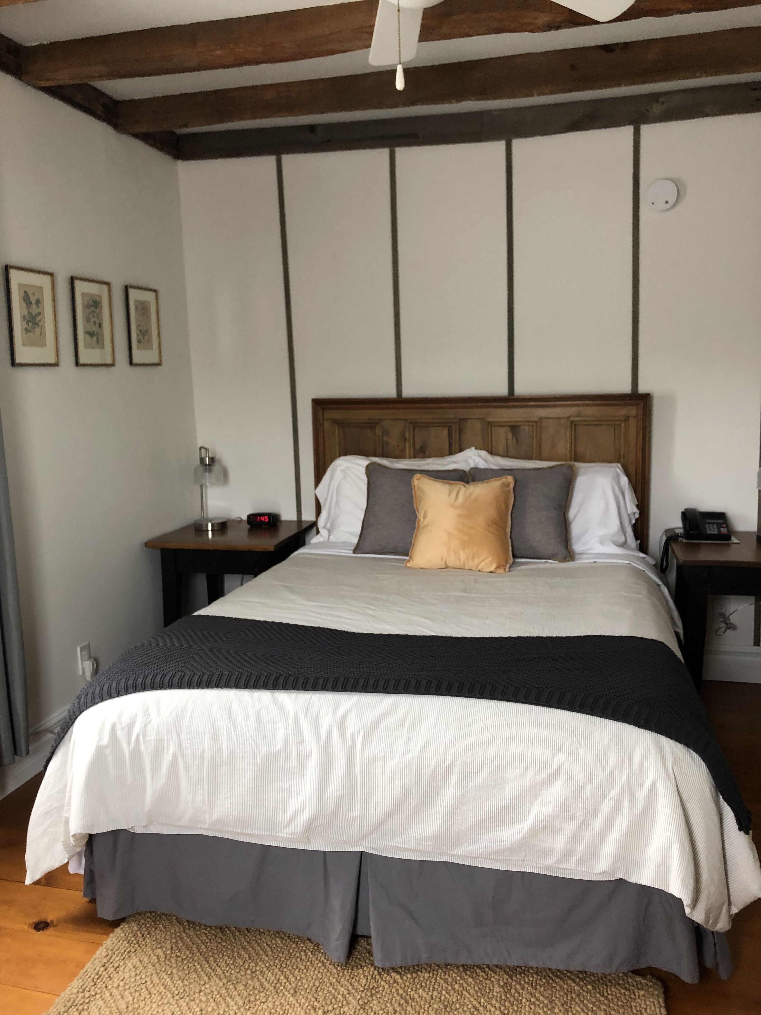 A bedroom with a wooden headboard, a large bed with a grey and white striped bedspread, and a beige throw blanket. There are bedside tables with lamps and phones on them.