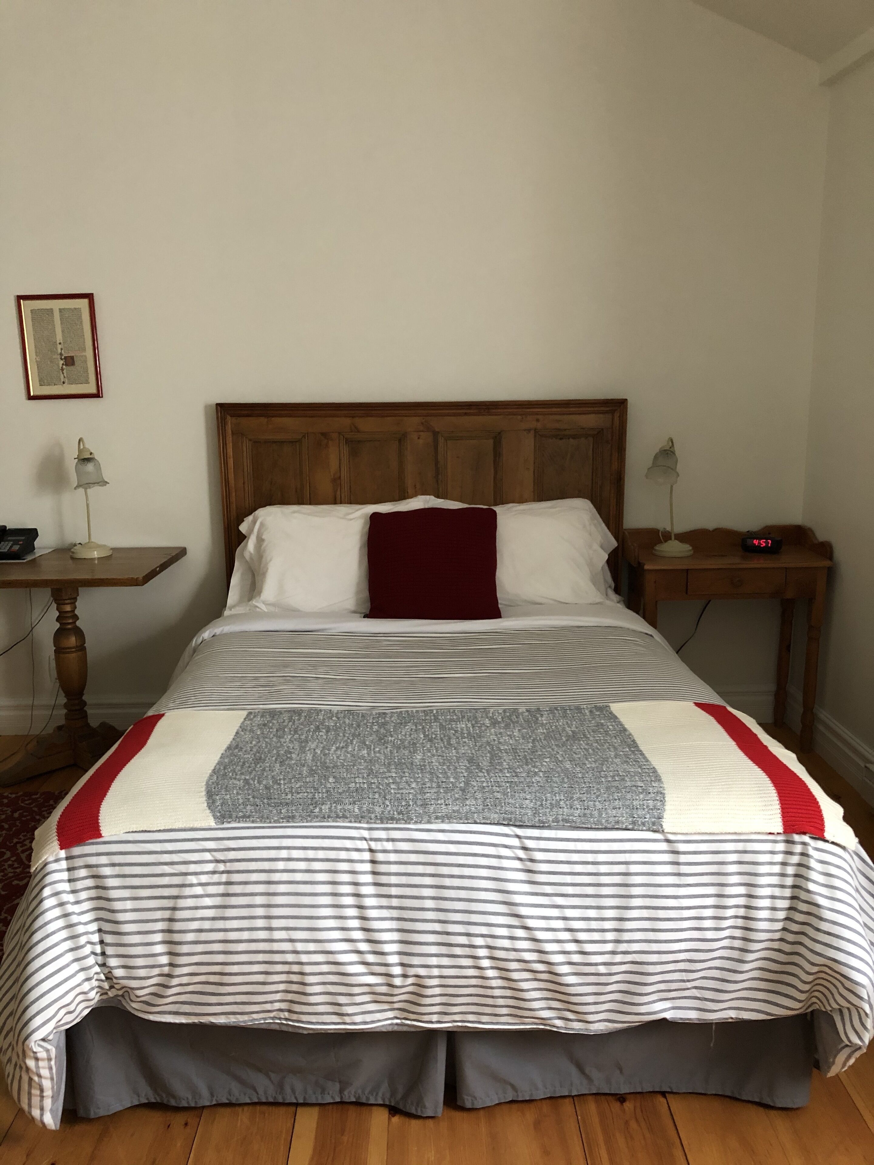 Una cama con cabecera de madera, almohada roja y colcha a rayas. Hay una mesita de noche con una lámpara y un reloj encima.