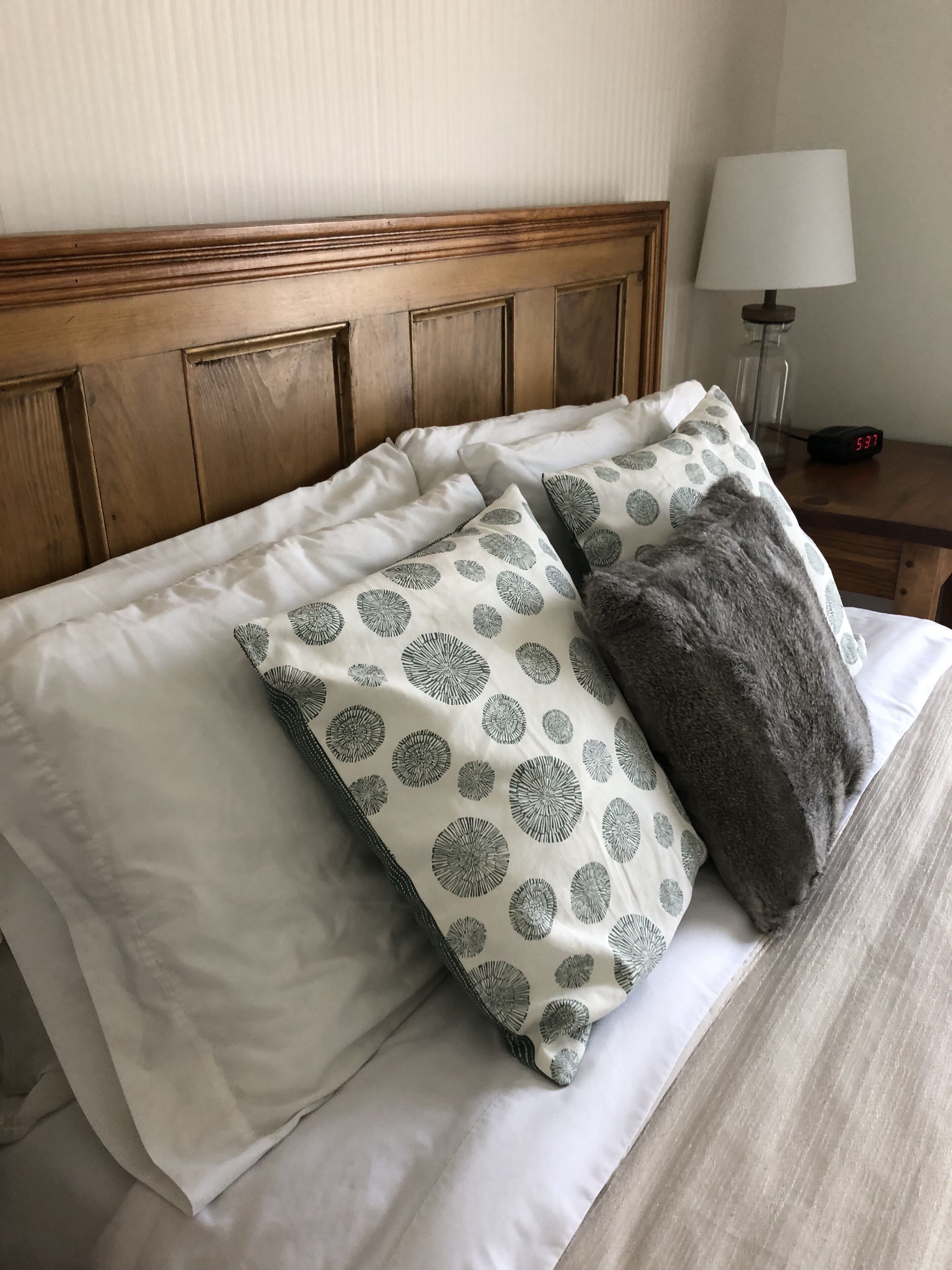 A neatly made bed with patterned pillows and a fluffy gray pillow, a wooden headboard, and a bedside table with a lamp and a digital clock.