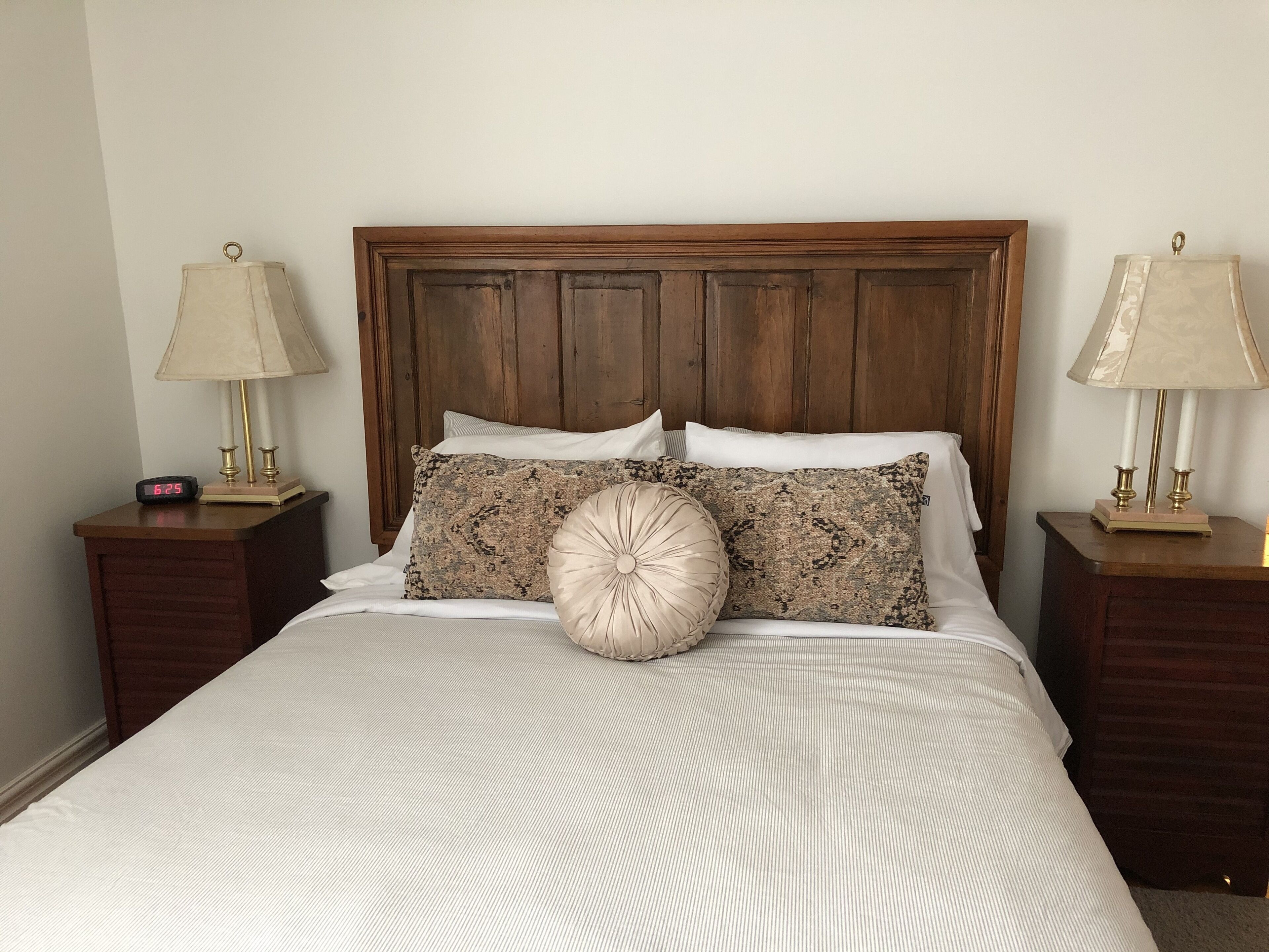 Una cama bien tendida con un cabecero de madera, dos mesitas de noche con lámparas y un reloj digital.