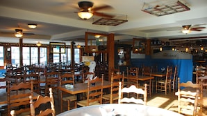 A restaurant with multiple wooden tables and chairs, ceiling fans, and large windows allowing natural light. - Auberge Knowlton (Lac-Brome)