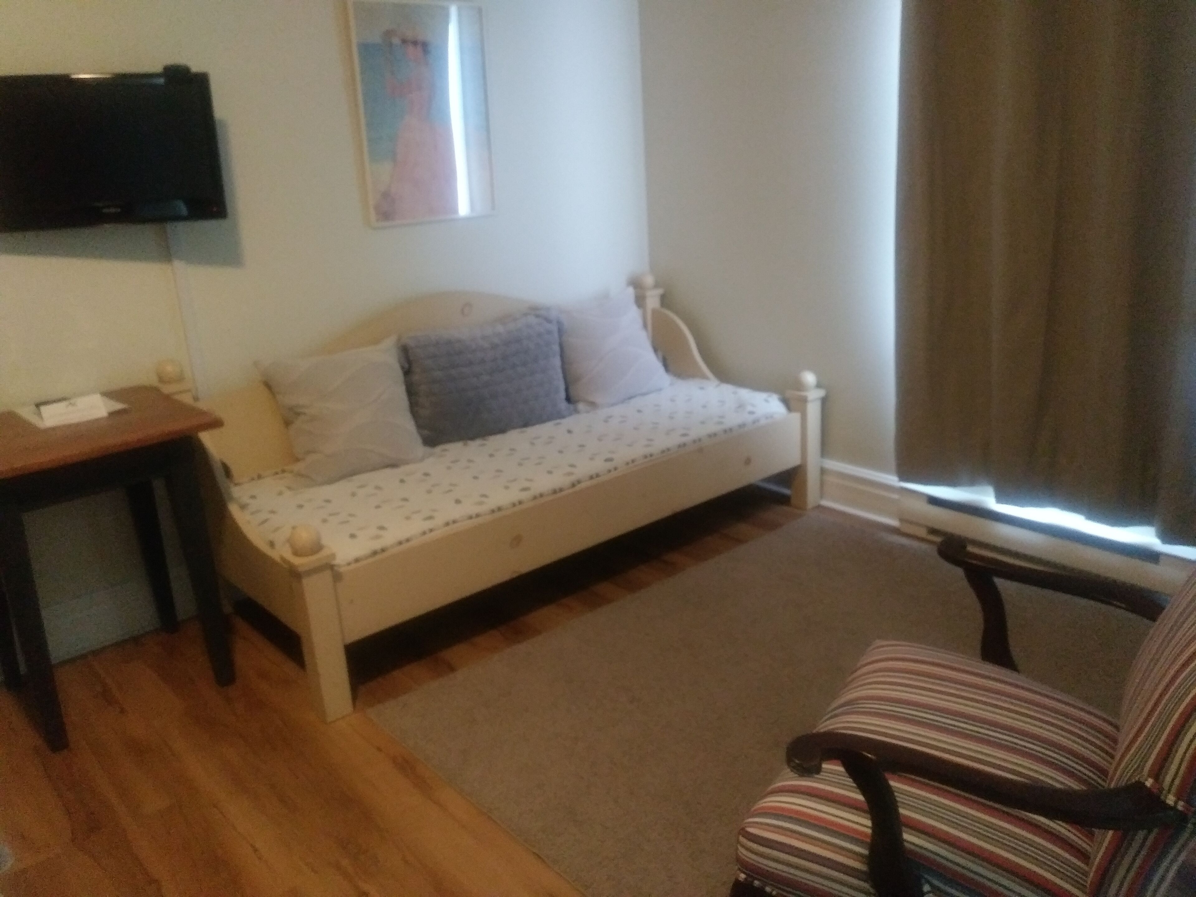 Una habitación pequeña y acogedora con una cama individual, una mesa de madera, una silla a rayas y un televisor montado en la pared.