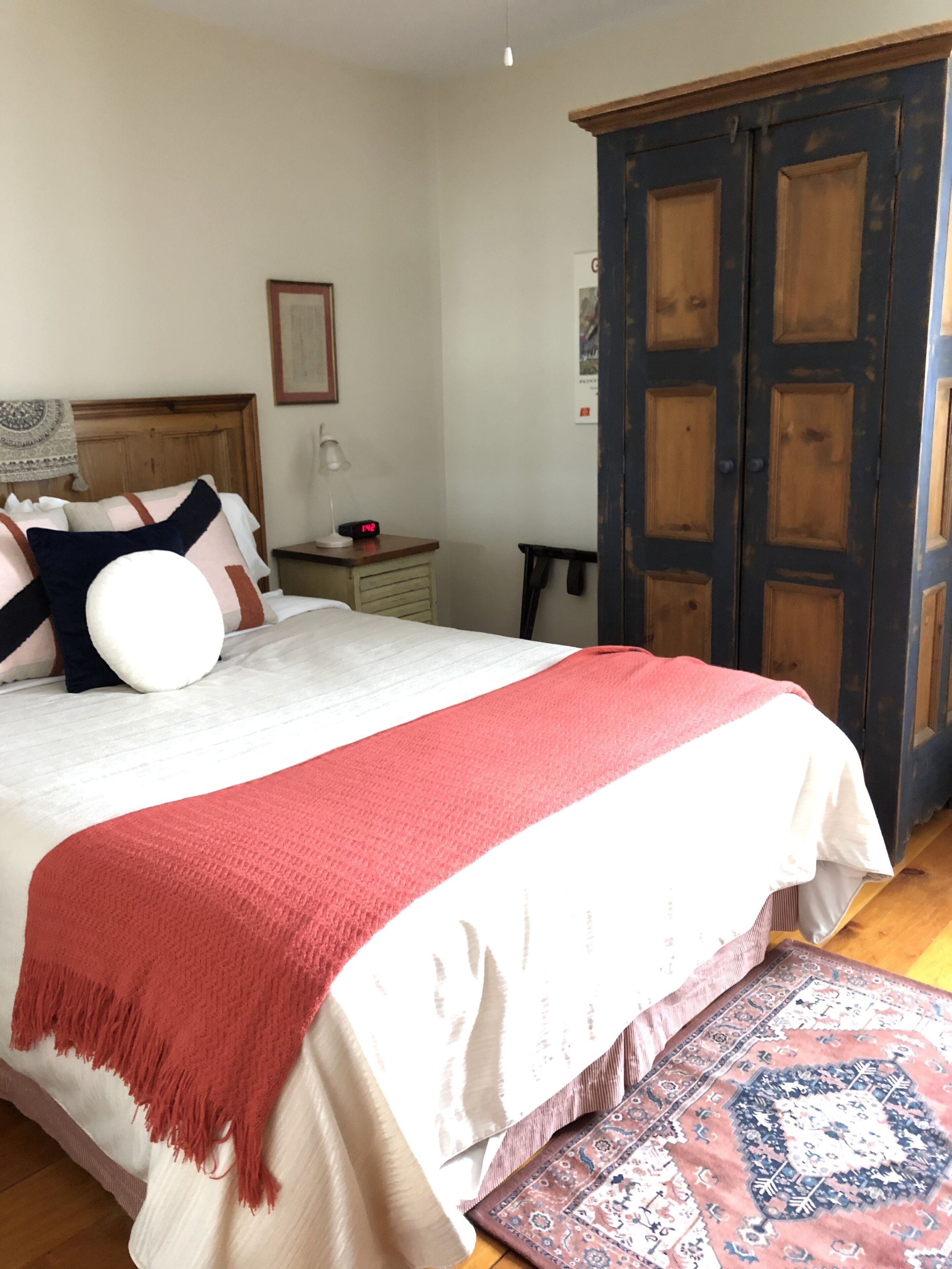 Un dormitorio con una cama, una mesita de noche, una lámpara, un reloj, un armario y una alfombra estampada.