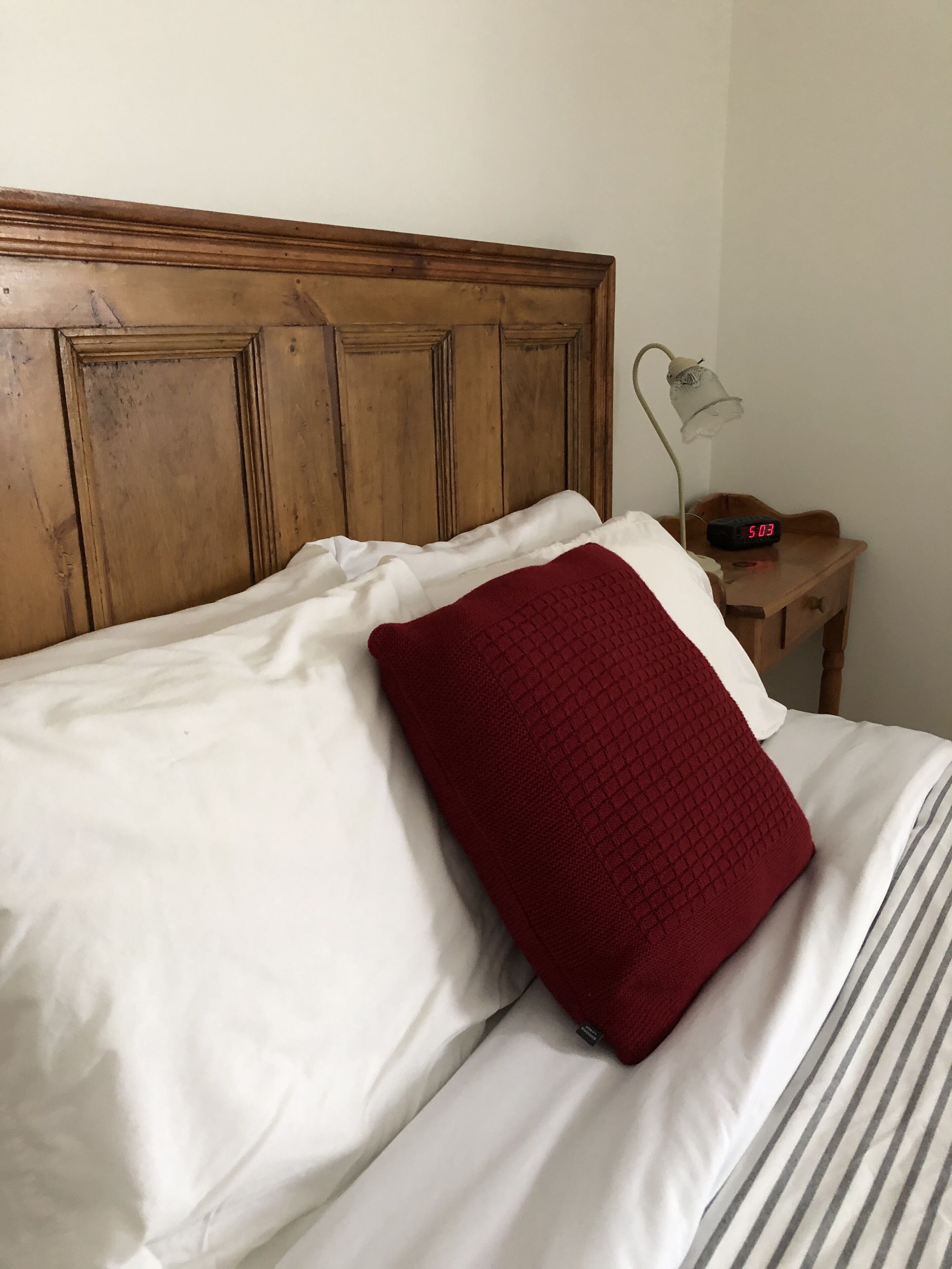 A bed with white linens and a red pillow, a wooden headboard, a bedside table with a lamp and a clock, and a striped blanket.