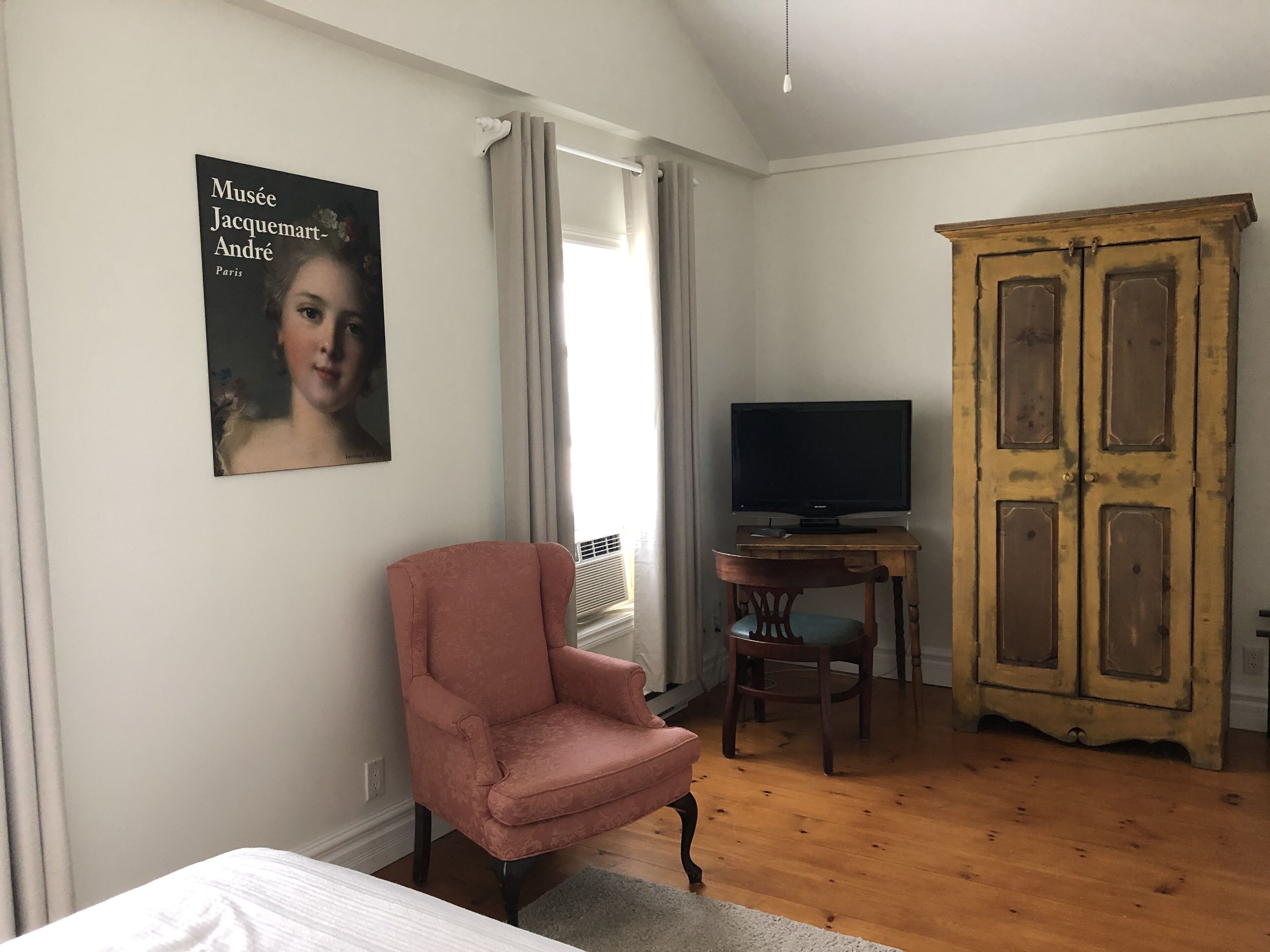Um quarto com uma cama, uma poltrona rosa, uma televisão sobre uma mesa de madeira, um guarda-roupa e um quadro na parede.