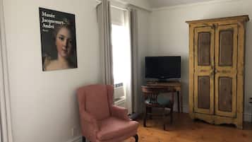 Um quarto com uma cama, uma poltrona rosa, uma televisão sobre uma mesa de madeira, um guarda-roupa e um quadro na parede.