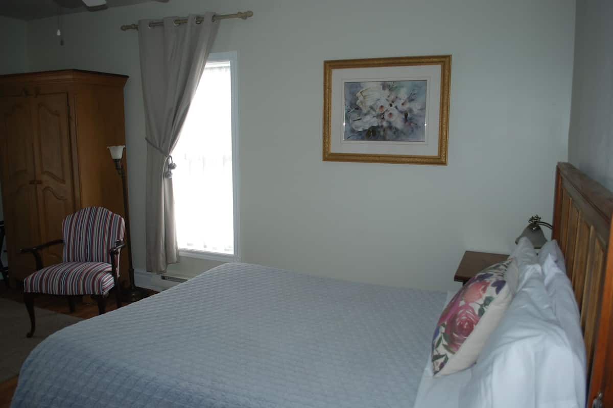 Un dormitorio con cama, una silla, armario, un cuadro en la pared y una ventana con cortinas.