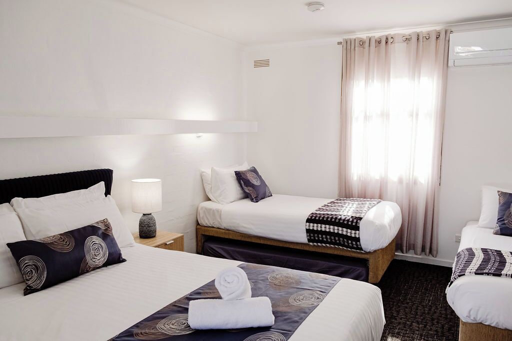 Habitación de hotel con dos camas, una mesita de noche, una lámpara y una ventana con cortinas.
