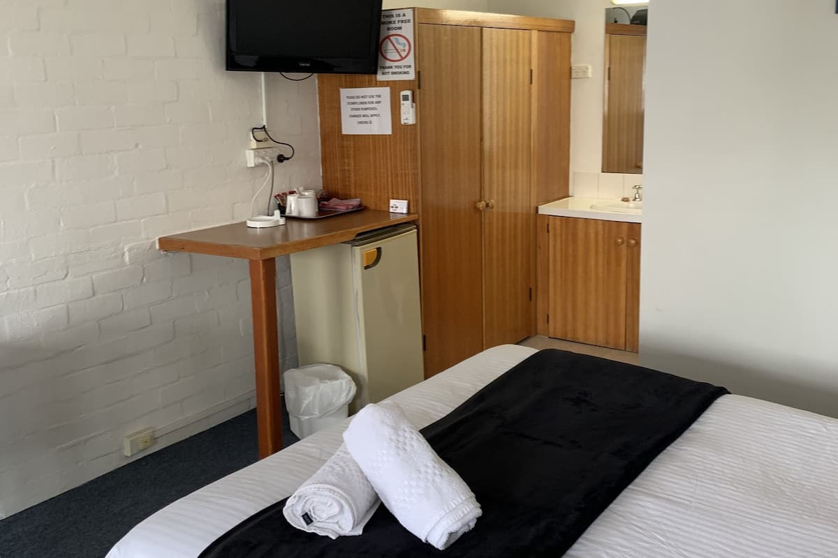 A hotel room with a bed, a TV mounted on the wall, a wooden wardrobe, a small desk with a mirror, and a trash bin.