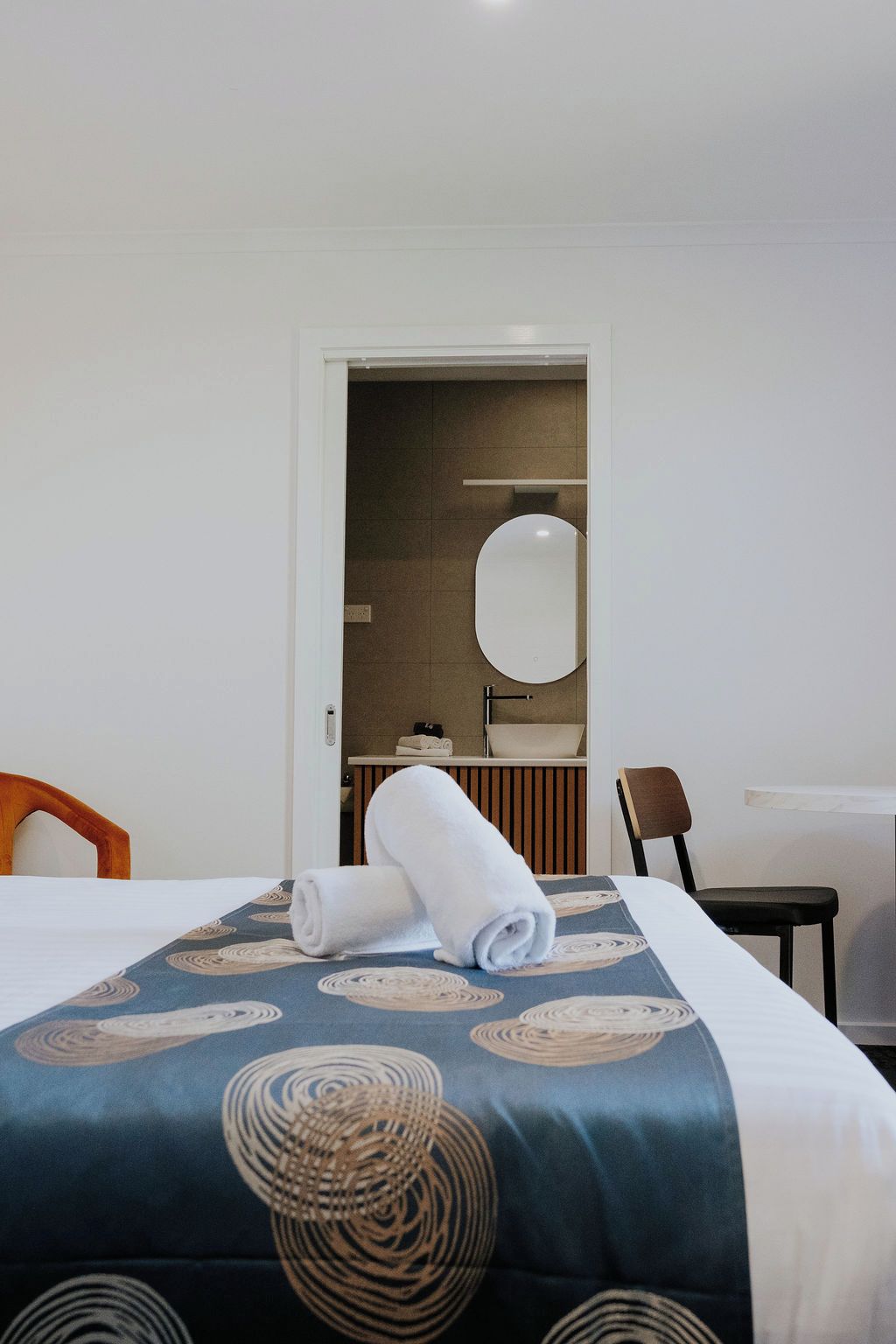 Habitación de hotel con cama, una silla, un escritorio, un espejo y un baño visible a través de una puerta abierta.