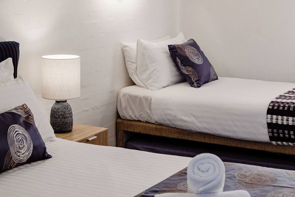 A hotel room with two beds, a nightstand with a lamp, and a wall-mounted light fixture.