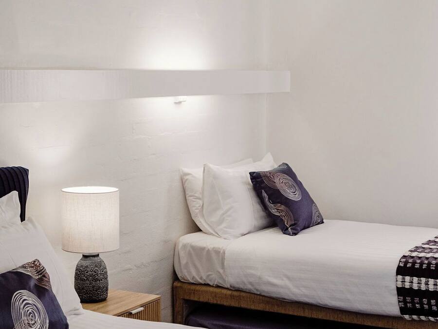 Hotellihuone, jossa on kaksi sänkyä, yöpöytä lampun kanssa ja seinään kiinnitetty valaisin.