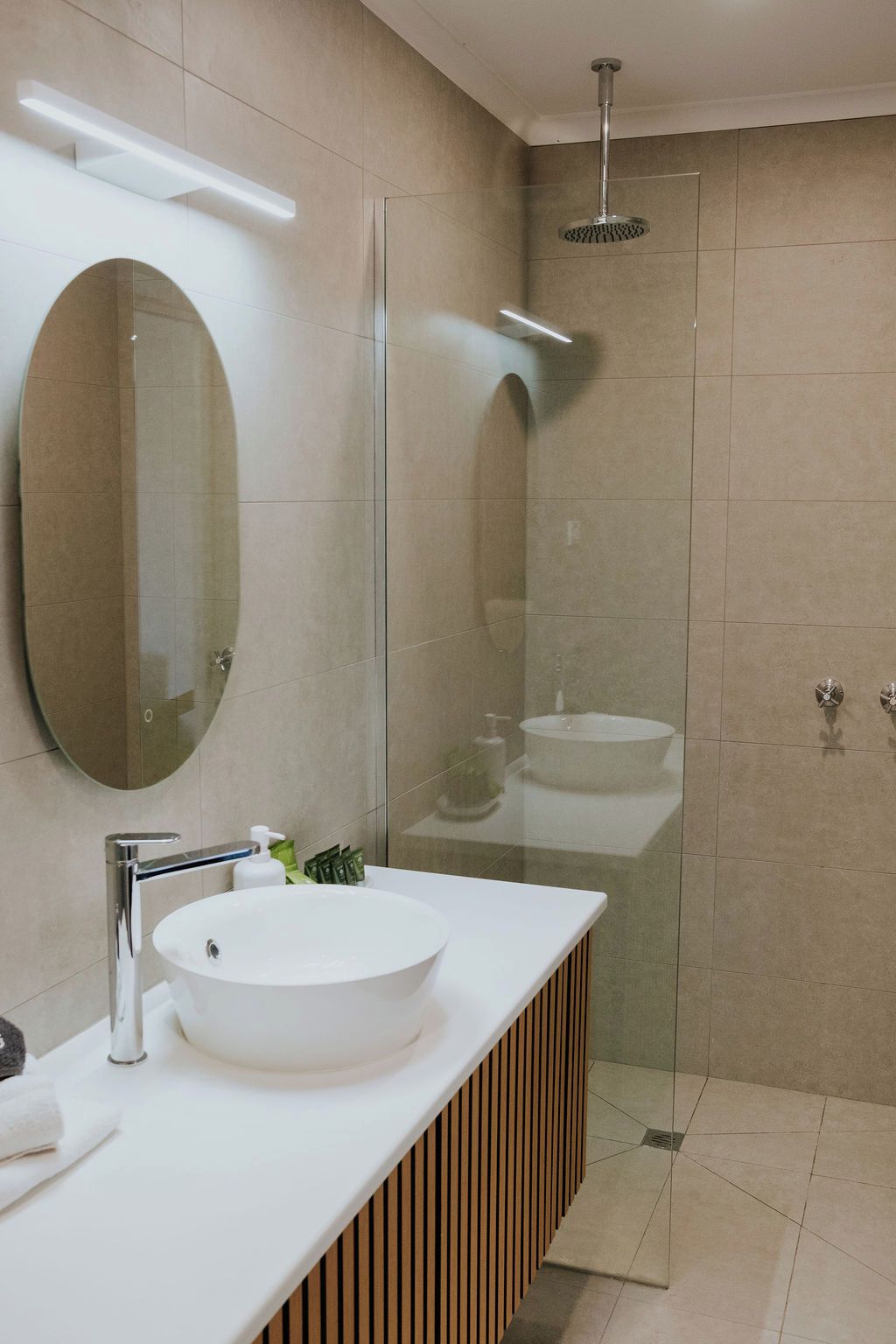 A modern bathroom with a large oval mirror, a round sink, and a glass-enclosed shower area.