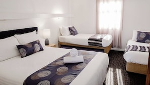 A hotel room with two single beds, a large bed, a bedside table with a lamp, a window with curtains, and a carpeted floor. - The Caledonian Inn (Port Fairy)